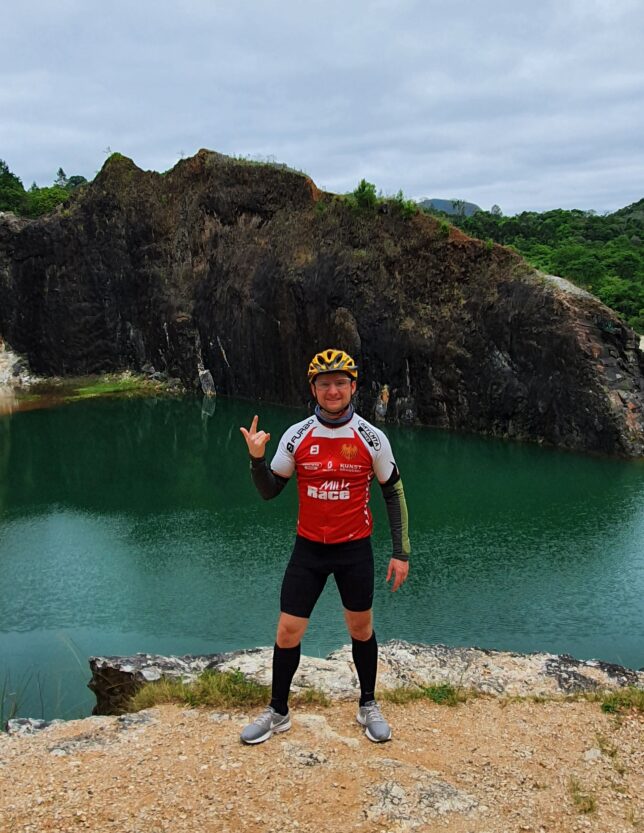 Eu (Daniel Scota), usando roupas adequadas para a prática do ciclismo, de capacete e óculos, em frente a Lagoa Azul, em Campo Magro.